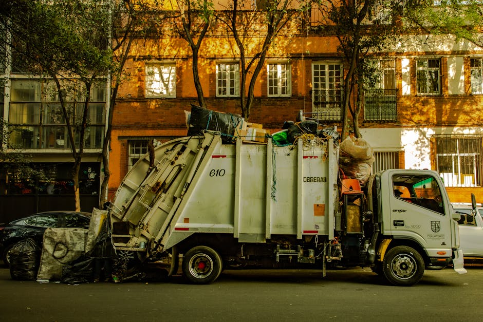 A white refuse collection truck for house waste is parked on a city street in front of a red-brick building with multiple windows. The truck's rear is open and filled with various garbage bags, cardbo