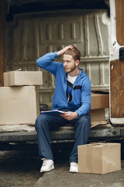 A man with a beard and wearing a black cap, white shirt, and jeans, standing in the back of a white moving van filled with packed cardboard boxes of various sizes, some wrapped in plastic and others e